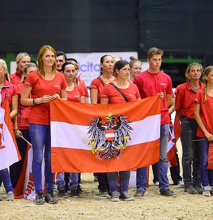 Eine Gruppe von Menschen in roten Shirts hält eine österreichische Flagge in einer Halle. Sie scheinen an einer Veranstaltung teilzunehmen.