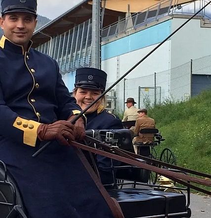 Persone in abiti storici guidano una carrozza trainata da cavalli davanti a un edificio moderno. Le uniformi sono blu con dettagli dorati.