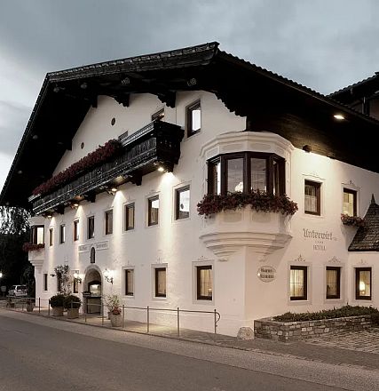 Ein traditionelles, alpenländisches Gebäude bei Dämmerung. Weiß verputzte Wände, braune Holzbalkone und Fensterläden, ruhige Straße im Vordergrund.