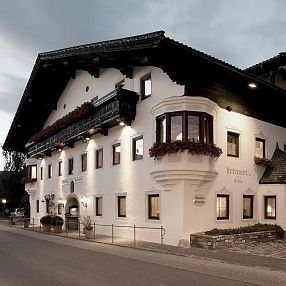 Un edificio tradizionale alpino bianco con finestre decorate e balconi in legno scuro, situato lungo una strada tranquilla, con il cielo nuvoloso sullo sfondo.