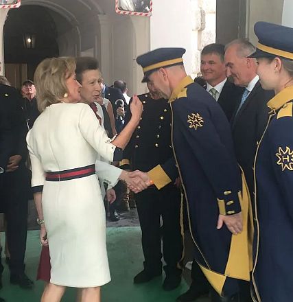 Eine Frau in einem weißen Kostüm schüttelt die Hand eines uniformierten Mannes, umgeben von mehreren Personen in formeller Kleidung.