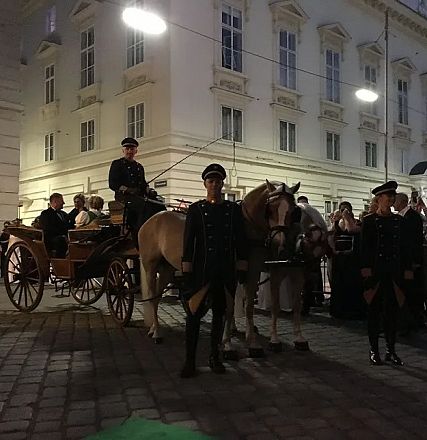 Eine Kutsche mit Pferden steht vor einem historischen Gebäude bei Nacht. Uniformierte Personen begleiten sie, während Menschen im Hintergrund zusehen.