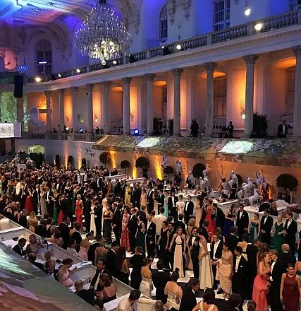 Elegante Veranstaltung in einem großen Ballsaal mit Gästen in Abendkleidung. Kronleuchter hängen von der Decke, Beleuchtung in warmen Farben.