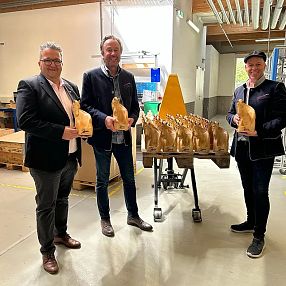 Three people in a factory setting, each holding a large loaf of bread. A table with more loaves is in the center. The room has industrial lighting and equipment.