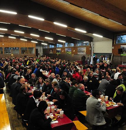 Grande sala con molte persone sedute a tavoli lunghi, in un ambiente rustico. Le persone chiacchierano e mangiano in un'atmosfera conviviale.