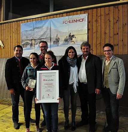 Un gruppo di persone in piedi sorride, con una donna al centro che tiene un certificato incorniciato. Sullo sfondo, un'immagine decorativa di cavalli.