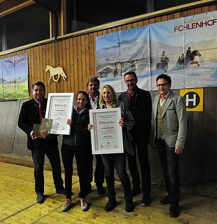Gruppo di persone in un edificio, due tengono dei certificati. Sfondo con immagini di cavalli e un logo giallo "H". Atmosfera formale e festosa.