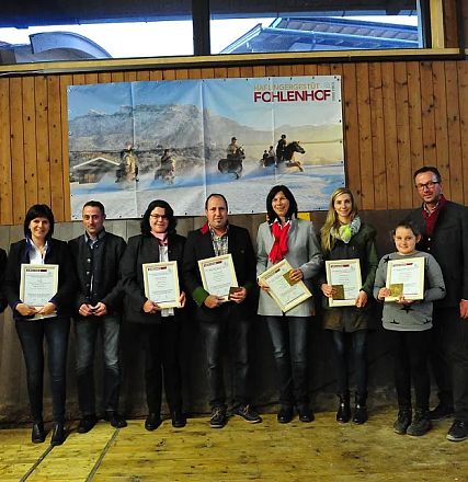 Gruppo di persone su un palco di legno con certificati. Sfondo di legno con poster di cavalli invernali. Atmosfera formale e sorrisi discreti.