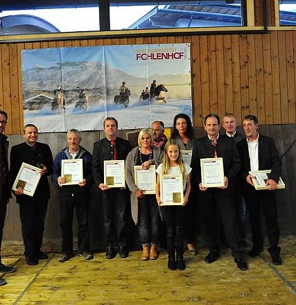 Un gruppo di persone in piedi su un palco in legno, ognuna che tiene un certificato. Sullo sfondo, un poster di cavalli e montagne.