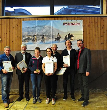 Un gruppo di persone in posa davanti a una parete di legno con certificati in mano. Al fondo, un poster di cavalli e montagne. Atmosfera di premiazione.