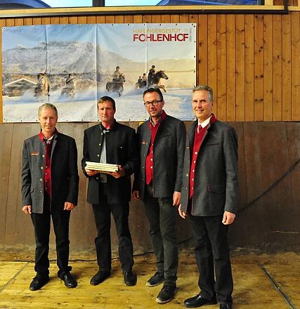 Quattro uomini in abiti eleganti posano davanti a un poster del "Fohlenhof" in un ambiente rustico con pannelli di legno e pavimento in legno.