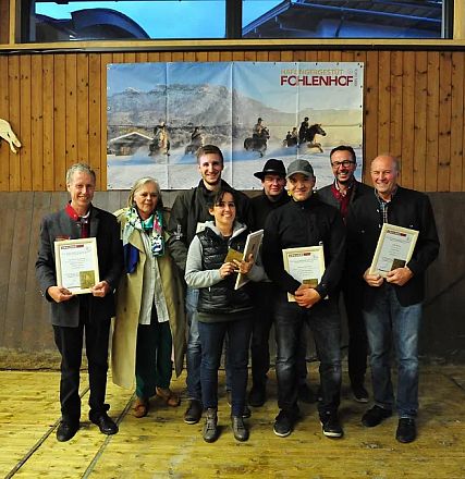 Foto di un gruppo di persone sorridenti che mostrano documenti e premi in un ambiente rustico con un'immagine di cavalli sullo sfondo.