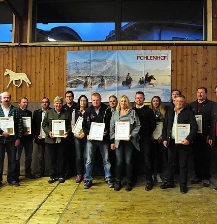 Un gruppo di persone posa su un palco di legno, tenendo certificati. Sullo sfondo, immagini di cavalli e un'insegna leggibile con "Pferdehof".