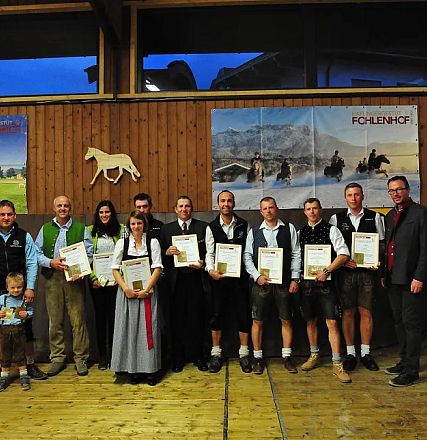 Una gruppo di persone in abiti tradizionali bavaresi posa per una foto di gruppo, tenendo certificati in una sala con pareti di legno e decorazioni equestri.