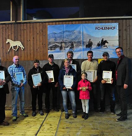 Un gruppo di persone in piedi in una sala, ciascuna con un certificato. Dietro di loro, una parete di legno con cartelloni di cavalli. Una bambina al centro.