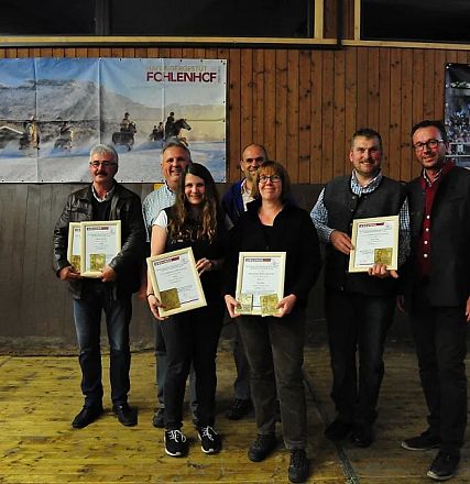 Un gruppo di persone sorridenti posa con certificati in una sala, con pannelli di legno e immagini di paesaggi alpini sullo sfondo.