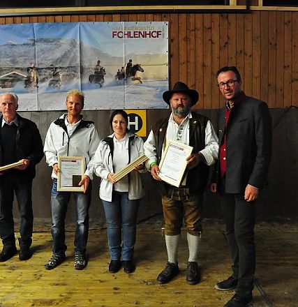 Cinque persone in piedi in una sala di legno, alcune con certificati in mano. Una parete con decorazioni e un cavallo stilizzato sullo sfondo.