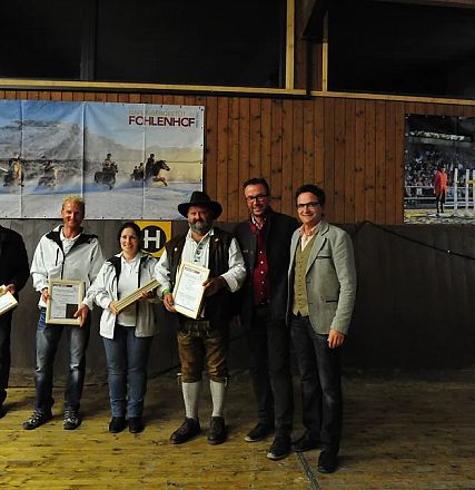 Un gruppo di sei persone posa insieme in una sala con pareti di legno. Due persone tengono certificati incorniciati. Due grandi poster sono appesi alla parete.
