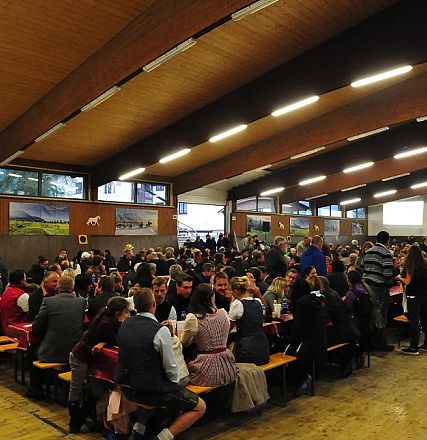 Una sala affollata con persone sedute a tavoli lunghi, tutti in abiti tradizionali. Il soffitto è in legno e ci sono grandi finestre ai lati.