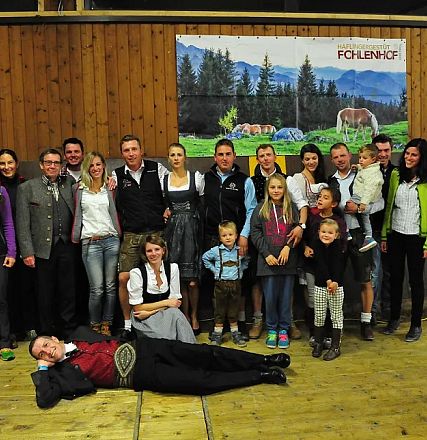 Gruppo di persone in abbigliamento tradizionale posano in una sala con pannelli in legno. Alcuni bambini sono in prima fila, mentre una persona è sdraiata a terra.