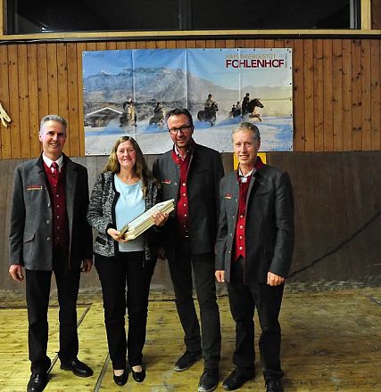 Quattro persone in piedi all'interno di una struttura in legno, dove una donna tiene un trofeo. Sullo sfondo, un poster con immagini di cavalli.