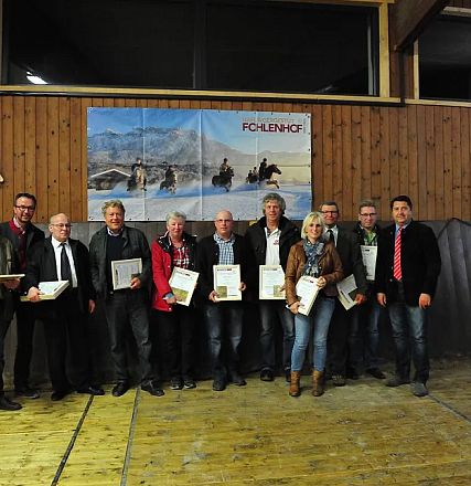 Un gruppo di persone posa per una foto in una sala interna, alcune tengono dei certificati. Sullo sfondo, poster di cavalli e un'insegna "Fohlenhof".