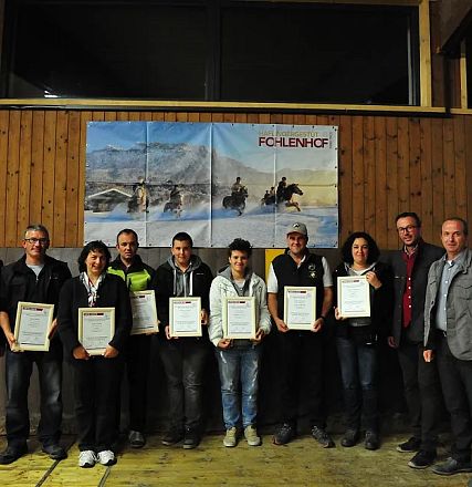 Un gruppo di persone in piedi in un ambiente interno, ognuna tiene un certificato. Dietro di loro, un pannello di legno con decorazione equina.