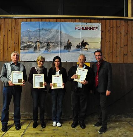 Cinque persone in piedi all'interno di un maneggio, ognuna con un certificato in mano. Una foto di cavalli e un'insegna "Fohlenhof" sullo sfondo.