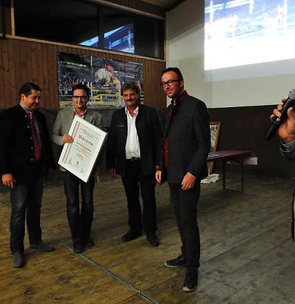 Un gruppo di uomini su un palco; uno tiene un grande documento incorniciato. Un altro parla al microfono mentre una presentazione scorre sullo schermo.