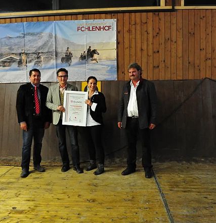 Quattro persone in abbigliamento formale posano con un certificato in un ambiente rustico. Sfondi con immagini di cavalli e sport equestri.