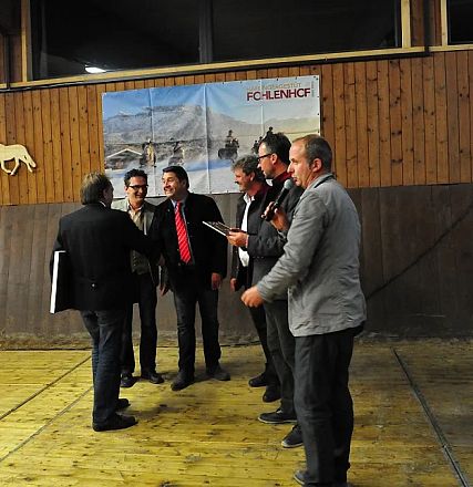 Gruppo di uomini in una sala, alcuni con fogli in mano. Pareti in legno, cartelloni di cavalli appesi. Atmosfera formale, forse una cerimonia.