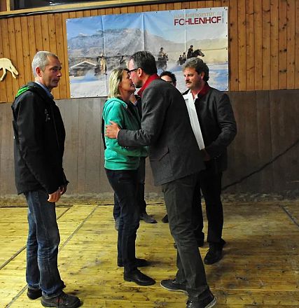 Persone si salutano in un ambiente rustico con pannelli di legno. Sullo sfondo ci sono poster di fattorie e cavalli.