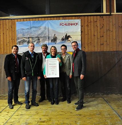 Sei persone in piedi su un palco di legno posano con un certificato in mano. Sullo sfondo, una parete di legno con un poster raffigurante cavalli.