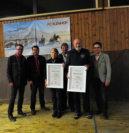Sei persone posano sorridenti davanti a un muro di legno con immagini e loghi. Due di loro tengono dei certificati incorniciati.