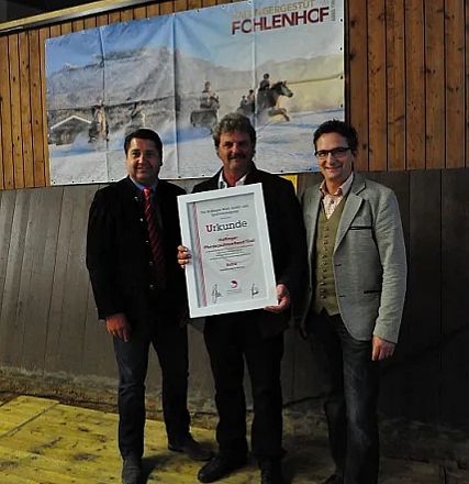 Tre uomini sorridenti posano con un grande certificato, apparentemente durante una cerimonia di premiazione. Sullo sfondo, un'immagine di cavalli in corsa.