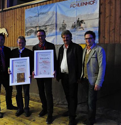 Cinque uomini in posa con due certificati incorniciati, indossano abiti formali, su un pavimento di legno, davanti a un poster con cavalli.