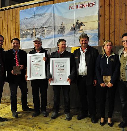 Un gruppo di persone posa con dei certificati in una stalla. Alcuni indossano abiti eleganti, il pannello dietro mostra immagini di cavalli.