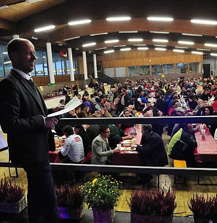 Un uomo parla a una folla seduta a tavoli lunghi in una grande sala. L'atmosfera è conviviale e vivace, con molte persone che mangiano e conversano.