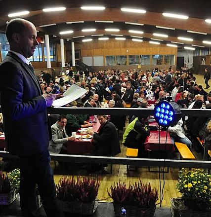 Uomo vestito formalmente parla a un pubblico numeroso seduto a tavoli lunghi in una sala illuminata, mentre tiene dei documenti in mano.