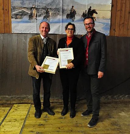 Tre persone sorridenti posano con certificati in mano, vestite in abiti formali, davanti a una parete di legno e un poster sullo sfondo.
