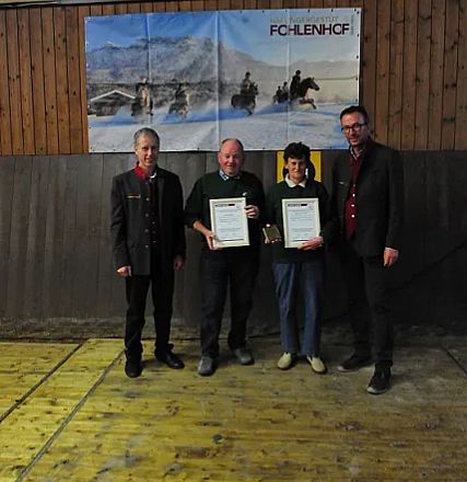 Quattro persone in piedi in un ambiente interno, due di loro tengono dei certificati in mano. Sullo sfondo, un pannello in legno e un'immagine appesa.