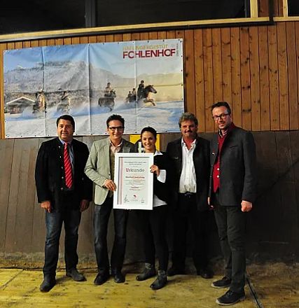 Sei persone in posa per una foto di gruppo in un ambiente interno con un banner e un certificato. L'atmosfera è formale e celebrativa.