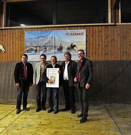 Cinque uomini in abiti formali posano davanti a un poster del "Fohlenhof" all'interno di un edificio in legno. Uno tiene una cornice con un certificato.