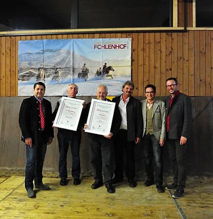 Sei uomini in posa sorridono davanti a un poster, due tengono dei certificati. L'ambiente ha pareti in legno e un'atmosfera formale e festosa.