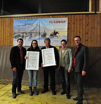 Cinque persone in piedi, sorridenti, con due di loro che tengono certificati incorniciati. Sullo sfondo, una parete di legno e un poster equestre.