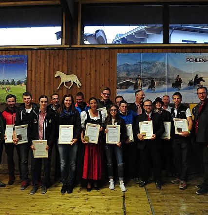 Un gruppo di persone sorridenti tiene in mano dei certificati in legno, posando per una foto in una sala con poster di cavalli alle pareti.
