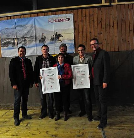 Sei persone sorridenti posano con certificati incorniciati davanti a una parete con legno e un poster di cavalli nel paesaggio invernale.