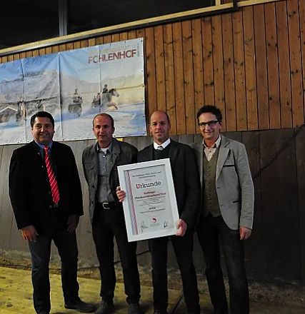 Quattro uomini sorridenti in abiti eleganti in posa con un certificato in una cornice bianca, davanti a uno sfondo di legno e un poster.