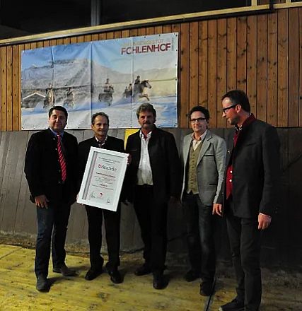 Cinque uomini in abiti formali posano insieme, uno tiene un grande certificato. Lo sfondo mostra un poster con cavalli e paesaggi innevati.