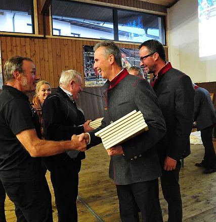 Un gruppo di persone in una sala, alcuni si stringono la mano. Un uomo tiene dei documenti. Ambiente informale, interazione amichevole.